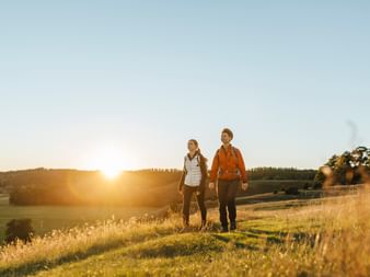 Two hikers with backpacks walking hand in hand across a sunlit meadow in Brösarp at sunset, with rolling fields and forests in the background.