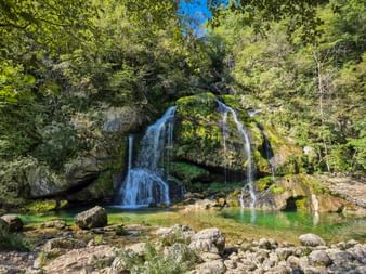 Virje-Wasserfall im Soča-Tal, der über moosbedeckte Felsen in ein klares türkisfarbenes Becken fließt, umgeben von üppigem Wald.