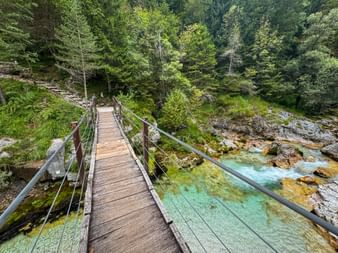 Holzsteg mit Seilgeländer über die türkisfarbene Soča in Trenta, umgeben von üppigem grünem Wald und felsigem Flussbett.