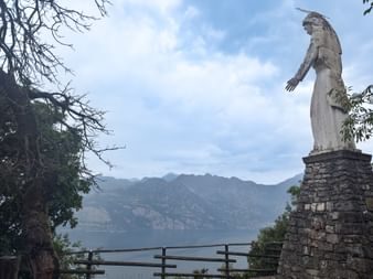 Steinerne Madonnenstatue auf einem Sockel mit ausgestrecktem Arm mit Blick auf den Gardasee, Berge im Hintergrund und Bäume rahmen die Szene.