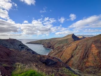 Dramatic volcanic cliffs in red and brown tones at São Lourenço Peninsula, Madeira, with blue ocean bay and two hikers visible on the ridge.