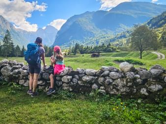 Zwei Wanderer mit Rucksäcken stehen an Steinmauer im Trenta-Tal, Julische Alpen. Grüne Wiesen und bewaldete Berge unter blauem Himmel.