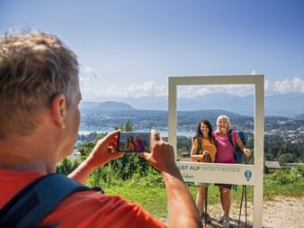 Mann fotografiert zwei Frauen am Velden-Fotorahmen mit See und Bergen im Hintergrund. Klarer blauer Himmel über Kärntner Landschaft.