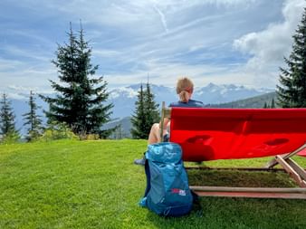 Wanderer mit blauem Rucksack sitzt auf roter Bank auf grüner Wiese mit Blick auf Bergpanorama mit schneebedeckten Gipfeln und Tannen im Pinzgau.