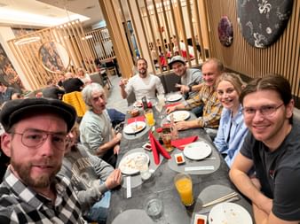 Gruppe von sieben Personen an einem Tisch in einem asiatischen Restaurant mit Holzlamellen, die gemeinsam eine Mahlzeit mit Tellern und Getränken genießen.