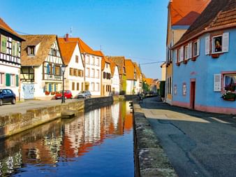 Schmaler Kanal durch eine elsässische Dorfstraße mit traditionellen Fachwerkhäusern mit orangen Ziegeldächern und bunten Fassaden.