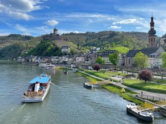 Passagierschiff auf der Mosel mit Uferpromenade, Kirchturm und Burg auf dem Hügel. Grüne Rasenflächen säumen das Ufer unter blauem Himmel.