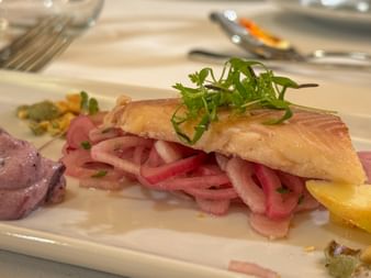 Pan-seared salmon fillet topped with fresh herbs, served on a bed of pink pickled onions with purple potatoes and lemon slices on a white plate.