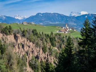 Ritten Earth Pyramids with tall rock spires surrounded by forest. Alpine village with church on green hills and snow-capped mountains behind.