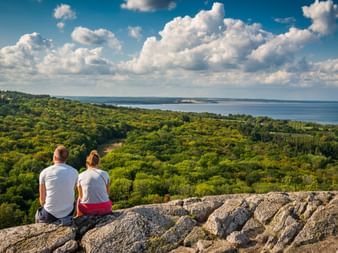 Zwei Wanderer sitzen auf felsigem Gipfel am Stenshuvud mit Blick auf grüne Wälder und blaue Ostseeküste unter bewölktem Himmel.