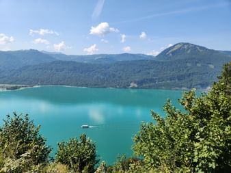 Panoramablick auf den türkisfarbenen Wolfgangsee im Salzkammergut mit bewaldeten Bergen, einem Passagierboot auf dem Wasser und blauem Himmel.