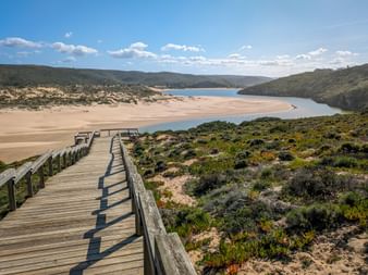 Wooden boardwalk winding through coastal vegetation on Rota Vicentina trail, overlooking a wide sandy beach and river estuary in Portugal.