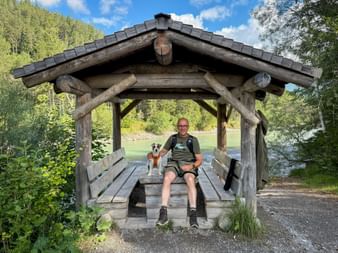 Hiker with glasses and small dog sitting in wooden shelter near Stanzach on the Lechweg trail. Green river and forest in background.