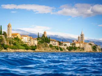 View from the sea of Rab Island's old town with four bell towers rising above stone buildings. Mountains visible in the background under blue sky.