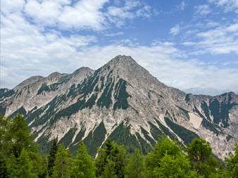 Mallestiger Mittagskogel mountain with rocky ridges and forested slopes under blue sky with white clouds in the Alpe-Adria region.