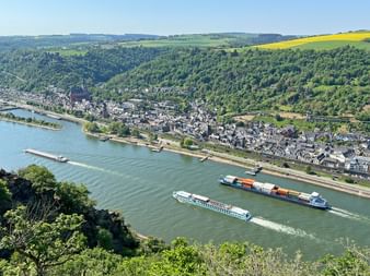 Luftaufnahme des Rheins mit Fracht- und Kreuzfahrtschiffen vor einer Uferstadt. Grüne Hügel und gelbe Felder im Hintergrund.