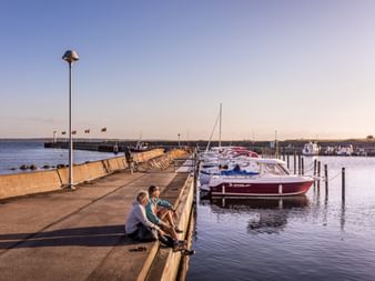 Zwei Personen sitzen auf einem Pier im Hafen von Viken und beobachten festgemachte Boote. Rote und weiße Motorboote liegen an Holzpfählen unter klarem Abendhimmel.