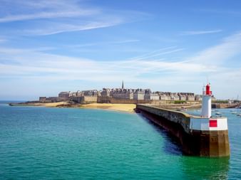 Rot-weißer Leuchtturm auf Steinpier im Hafen von St. Malo. Türkisfarbenes Wasser, Sandstrand und historische Stadtmauer im Hintergrund.
