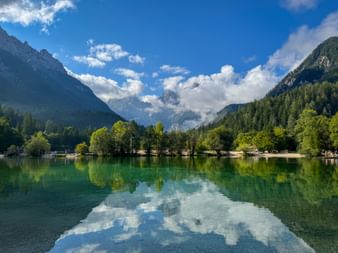 Jasna See mit türkisfarbenem Wasser, das Wolken und Himmel spiegelt, umgeben von bewaldeten Ufern und Julischen Alpen im Hintergrund.