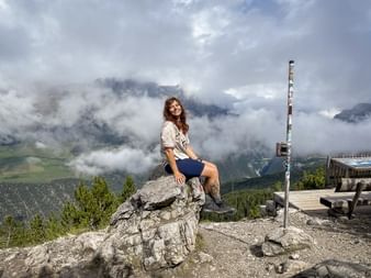 Woman Anna-Lu sitting on rock at mountain summit with hiking boots, cloudy Alpine peaks and green valleys visible behind her.
