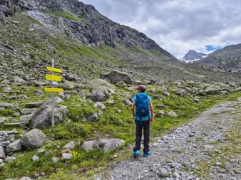 Wanderin mit blauem Rucksack steht an gelben Wegweisern in felsigem Alpental bei Neukirchen, Pinzgau, mit Bergen unter bewölktem Himmel.