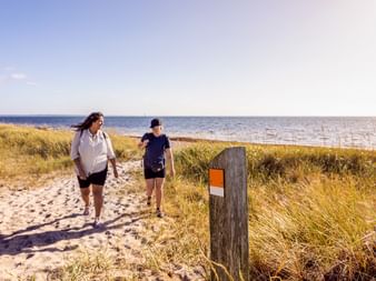 Zwei Wanderer auf sandigem Küstenpfad mit Wegmarkierung, Dünengras und Meerblick unter klarem Himmel in Schweden.