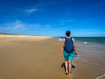 Female hiker with blue backpack walking barefoot along sandy beach with turquoise ocean waves under clear blue sky on Portuguese Camino.