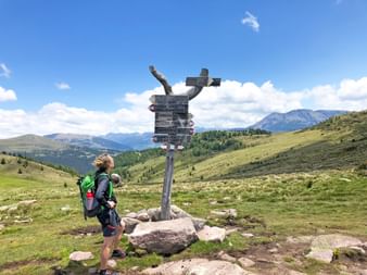 Hiker with backpack standing at wooden directional signpost on Möltern circular trail in South Tyrol, with green alpine meadows and mountains.