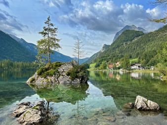 Crystal clear Hintersee lake with rocky island and trees, surrounded by forested mountains in the Bavarian Alps. Buildings visible on shoreline.