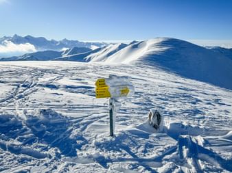 Gelber Wanderwegweiser im Schnee auf einem verschneiten Bergplateau in Maria Alm-Dienten, mit schneebedeckten Gipfeln und blauem Himmel.