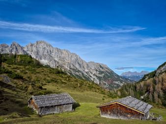 Two traditional wooden alpine huts on green meadows in Grieseltal valley with rocky mountain peaks and blue sky in the Kitzbühel Alps.