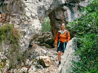 Smiling hiker in orange jacket walking on rocky trail surrounded by limestone cliffs and green vegetation on Sardinia's east coast.