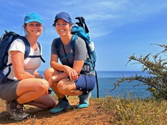 Two smiling female hikers with backpacks crouching on a coastal trail in the Algarve, with blue ocean and sky in the background.