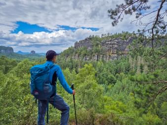 Hiker with blue backpack and trekking pole viewing sandstone cliffs and forest from Affensteinpromenade on the Malerweg trail.