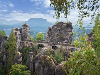 Historische Basteibrücke zwischen hoch aufragenden Sandsteinfelsen, eingerahmt von grünem Laub mit fernen Bergen im Hintergrund.
