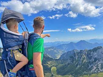 Mann im grünen Shirt mit Kind in blauer Rückentrage zeigt auf Bergpanorama in Bayern. Grüne Täler und felsige Gipfel unter blauem Himmel.