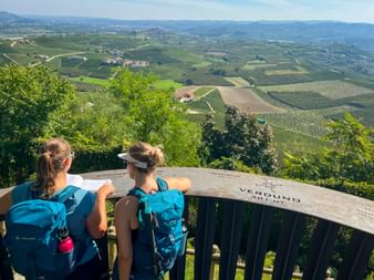 Two hikers with backpacks at Verduno viewpoint overlooking rolling hills, vineyards, and villages in Piedmont-Liguria region.