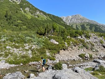 Wanderer mit blauem Rucksack watet durch flachen Bergbach am Lechweg. Grüne Hänge und felsige Gipfel unter blauem Himmel im Hintergrund.