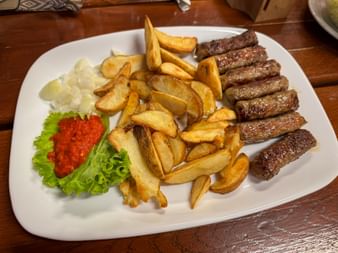 Weißer Teller mit gegrillten Ćevapčići-Würstchen, goldenen Pommes, Zwiebeln, roter Ajvar-Sauce und Salat auf Holztisch in Trenta, Julische Alpen.
