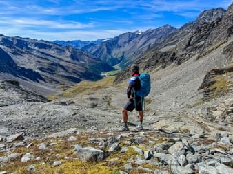Wanderer mit blauem Rucksack auf felsigem Bergpfad mit Blick auf das Pfossental. Alpine Gipfel und grünes Tal in der Ferne unter blauem Himmel.
