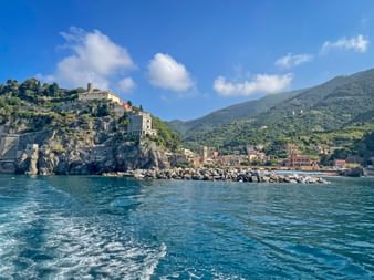 Monterosso al Mare village in Cinque Terre with colorful buildings along the coast, rocky cliffs, a stone breakwater, and green mountains under blue sky.