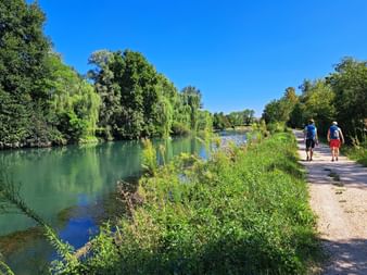 Zwei Wanderer auf Kiesweg neben türkisfarbenem Sile-Fluss mit üppiger grüner Vegetation und Trauerweiden unter klarem blauen Himmel.