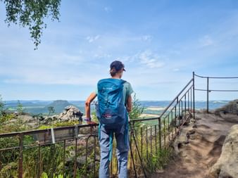 Hiker with blue backpack at Papststein viewpoint on Malerweg, overlooking forested valleys and distant rock formations under blue sky.