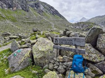 Hölzerner Wegweiser zum Nationalpark Hohe Tauern und Kernzone zwischen Felsen mit blauem Rucksack. Berglandschaft mit grünen Hängen.