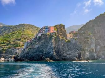 Colorful houses of Manarola perched on dramatic rocky cliffs above turquoise sea water. Terraced vineyards cover the hillside under blue sky.