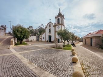 White Igreja de Santiago church with bell tower in Castelo de Neiva. Stone-paved square with spherical bollards and olive trees under cloudy sky.