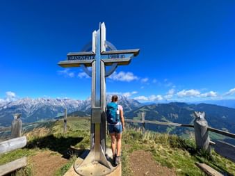 Female hiker with backpack standing at metal summit cross marked Klingspitz 1988m, overlooking Alpine mountain ranges under blue sky with clouds.