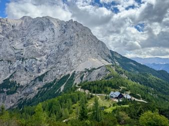 Berghütte auf einem bewaldeten Hügel am Vršič-Pass in den Julischen Alpen, mit dramatischen Felsgipfeln und grünen Wäldern unter bewölktem Himmel.
