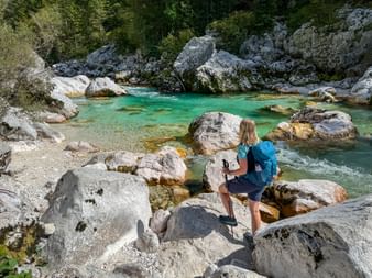 Wanderin mit blauem Rucksack sitzt auf weißen Kalksteinfelsen am türkisfarbenen Soča-Fluss in den Julischen Alpen, umgeben von Wald.