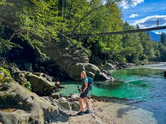 Wanderin mit Rucksack am felsigen Ufer der türkisfarbenen Soča. Eine Hängebrücke überspannt den Fluss, umgeben von Wald.